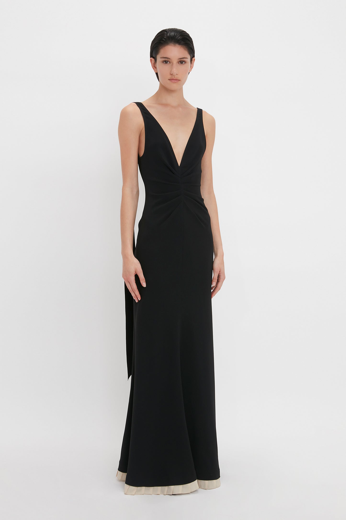 Person posing in a sleeveless, floor-length V-Neck Gathered Waist Floor-Length Gown In Black by Victoria Beckham against a plain white background, accentuating a deep-V neckline and flattering silhouette.