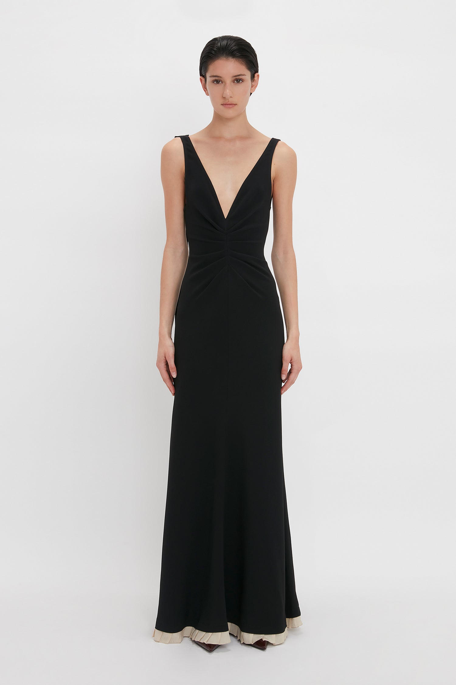 A person stands against a plain background wearing a Victoria Beckham V-Neck Gathered Waist Floor-Length Gown In Black with a deep V-neckline, slightly flared at the hem, creating a flattering silhouette.