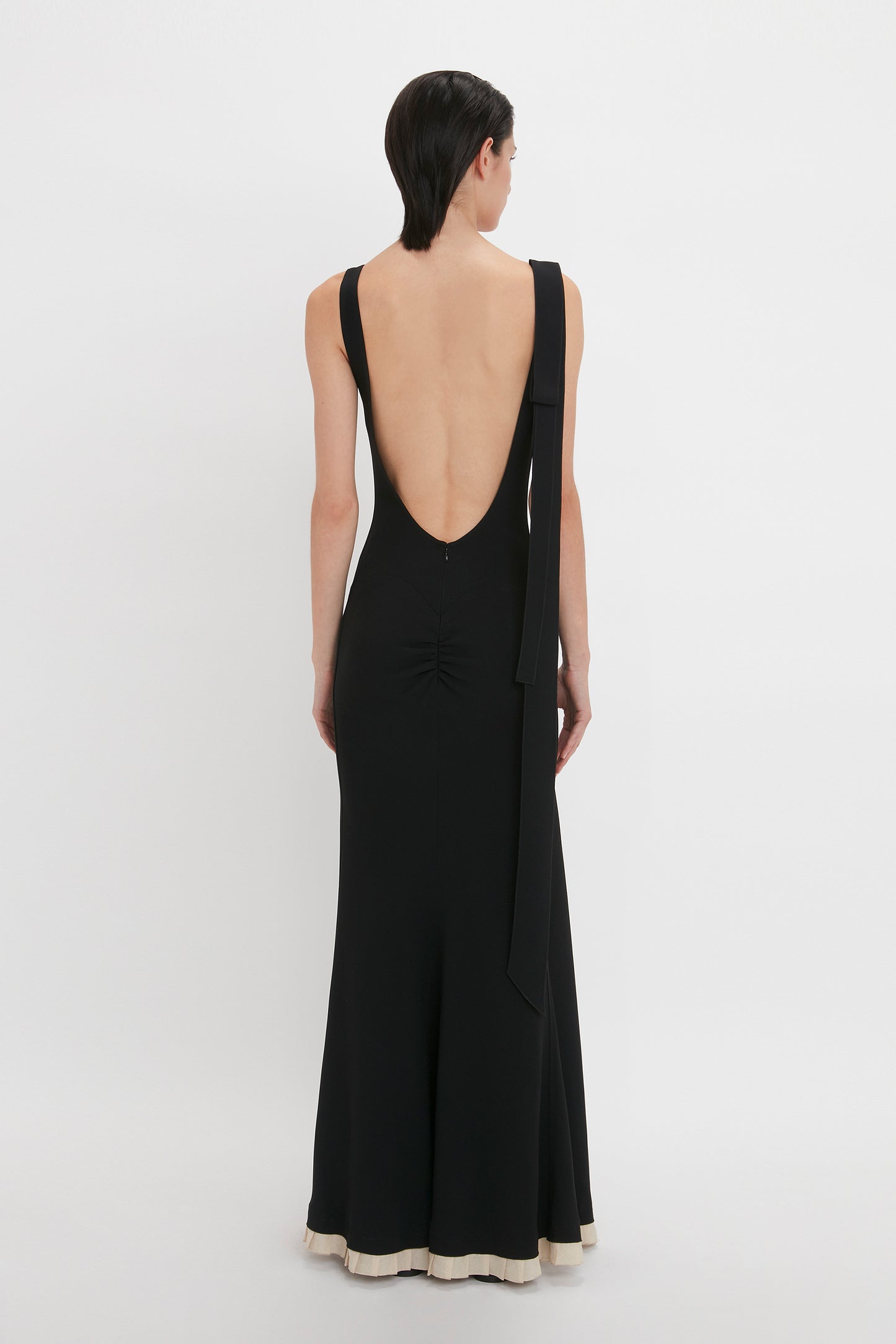 Person wearing a black, backless V-Neck Gathered Waist Floor-Length Gown In Black by Victoria Beckham with white trim at the hem, featuring a deep-V neckline and a flattering silhouette, standing against a plain white background.
