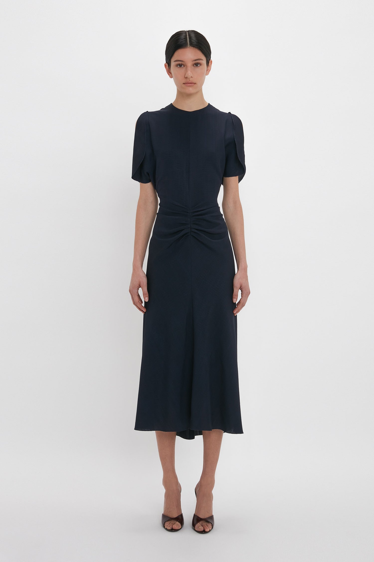 A woman stands against a plain white background, wearing the Gathered V-Neck Midi Dress In Midnight by Victoria Beckham, paired with brown open-toe heels.