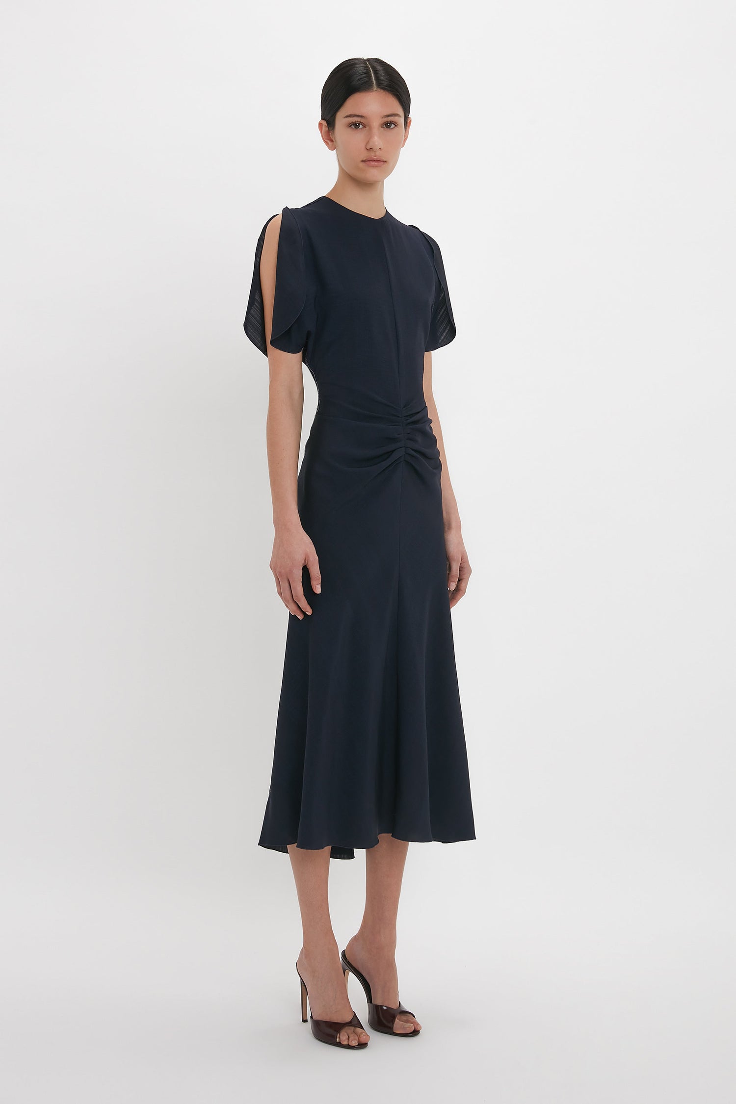 A person in a Victoria Beckham Gathered V-Neck Midi Dress In Midnight stands facing the camera, hands relaxed by their sides, and wearing black heels.