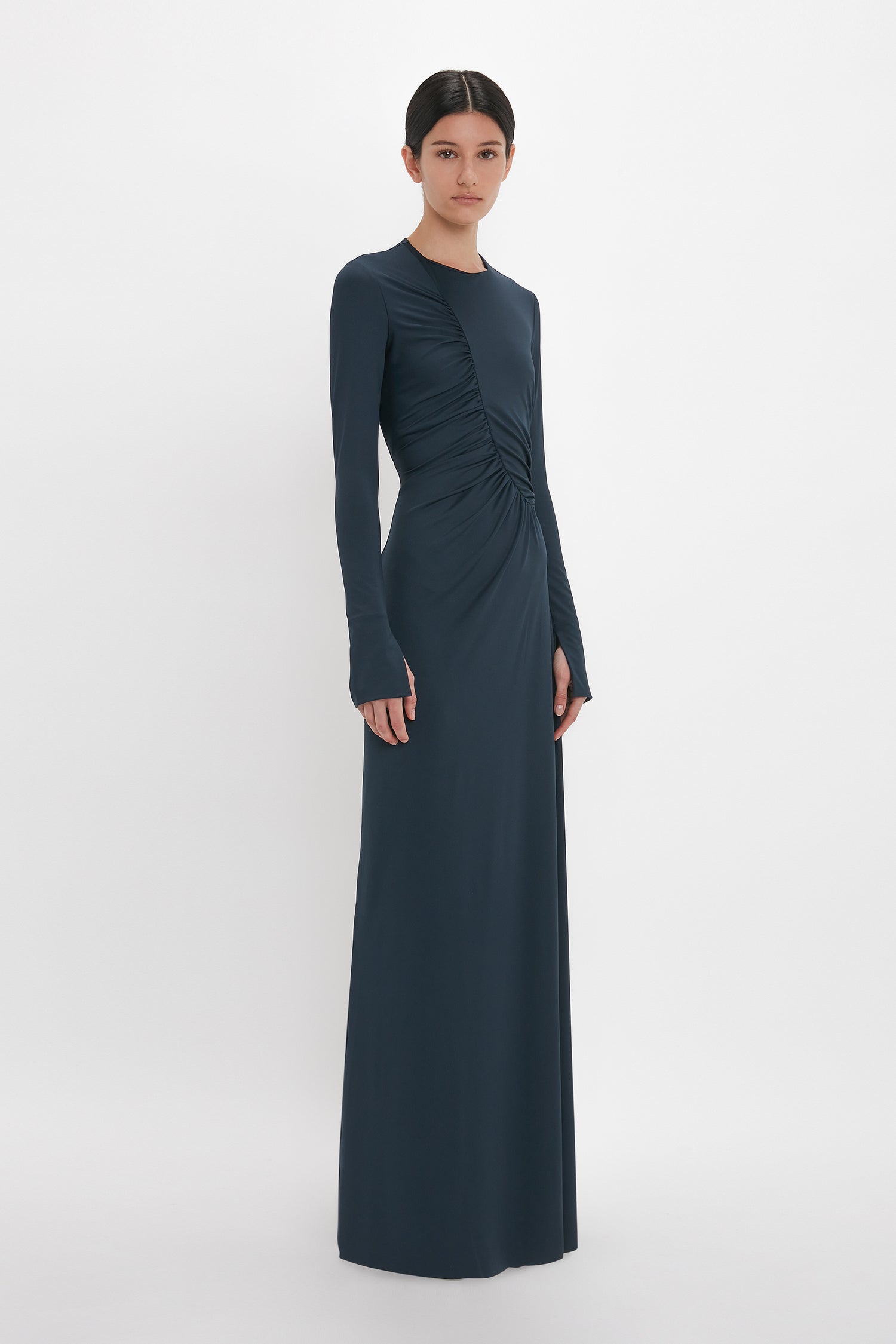 A woman stands wearing a dark blue, long-sleeve, floor-length dress with ruched detailing on the front. Her elegant Ruched Detail Floor-Length Gown In Midnight by Victoria Beckham is posed against a plain white background.