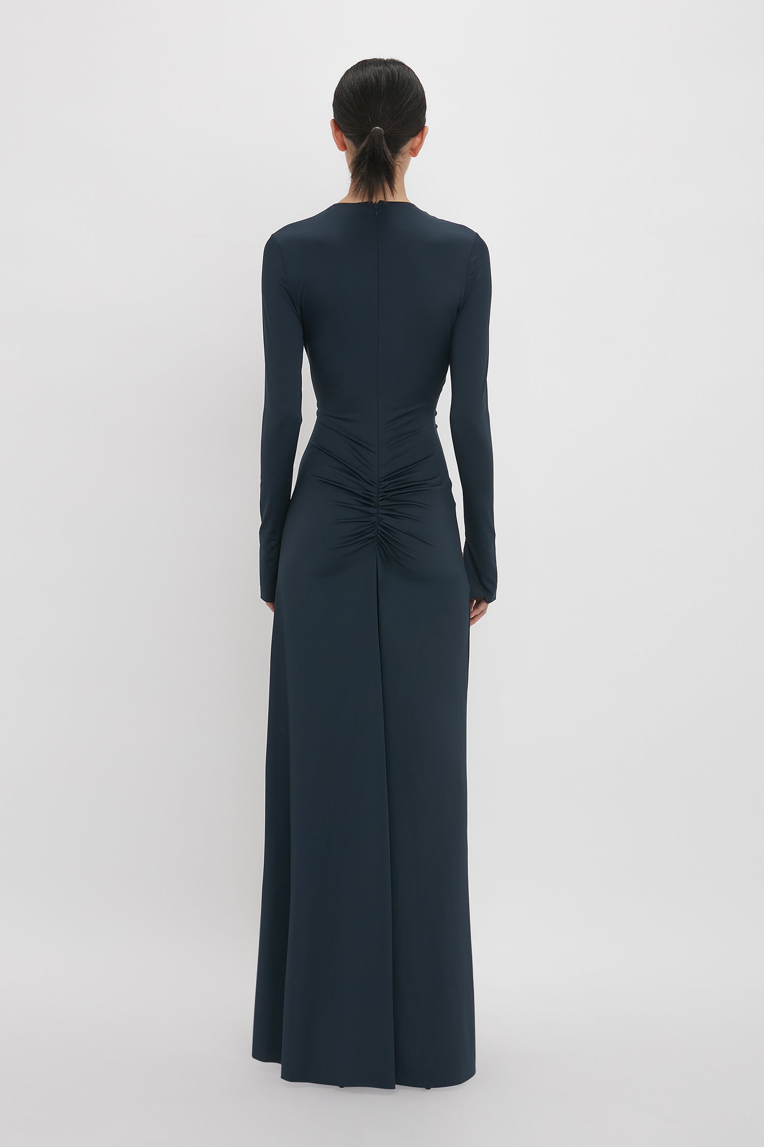 A person with dark hair tied in a low ponytail is wearing a long-sleeved, floor-length Ruched Detail Floor-Length Gown In Midnight by Victoria Beckham seen from the back against a plain white background.