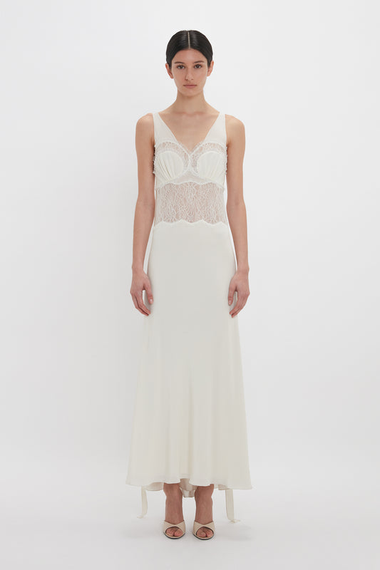 A person stands against a plain white background, wearing the Victoria Beckham Ruffle Detail Midi Dress In Ivory featuring sheer tactile lace details and a long, flowing skirt. They have dark hair pulled back and are wearing strappy heels.