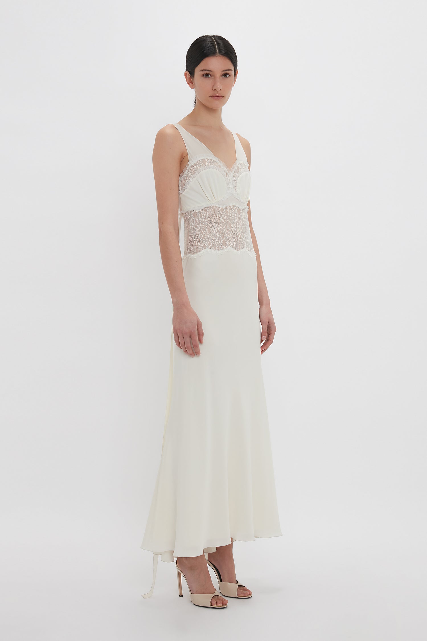 A person wearing the Ruffle Detail Midi Dress In Ivory by Victoria Beckham, with tactile lace detailing on the bodice and thin spaghetti straps, stands against a plain white background.