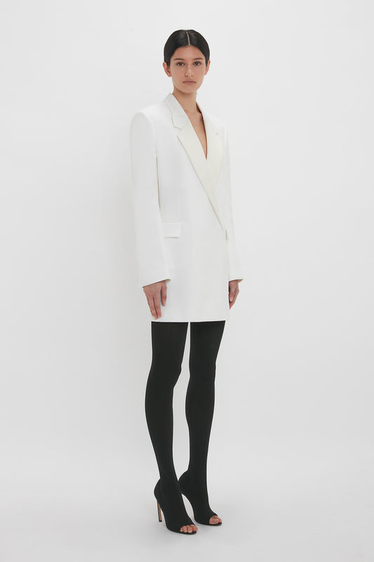 A woman dressed in a stylish double-breasted Exclusive Fold Shoulder Detail Dress In Ivory by Victoria Beckham and black tights stands against a white background, looking directly at the camera.