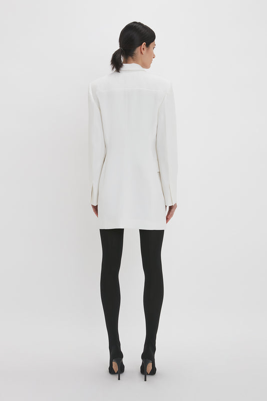 A woman viewed from behind, wearing a Victoria Beckham Exclusive Fold Shoulder Detail Dress In Ivory and black tights with high heels, posing against a plain white background.
