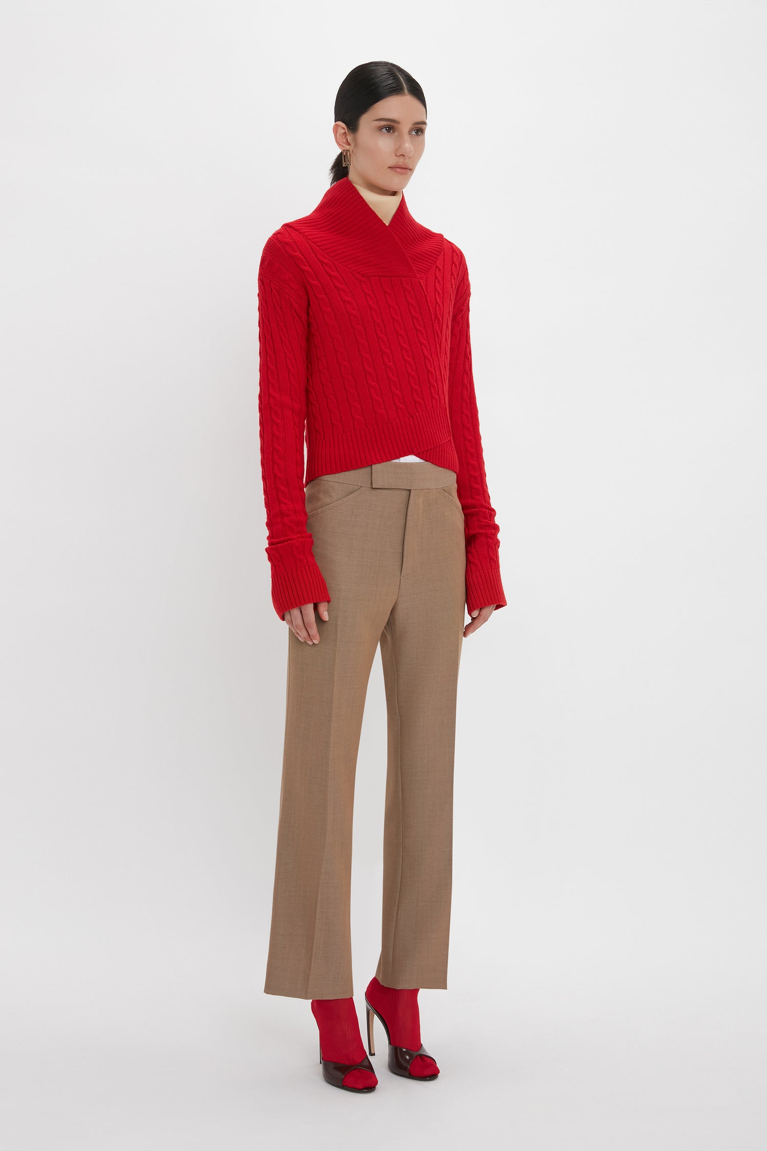 A person is wearing a luxurious red Wrap Detail Jumper In Red by Victoria Beckham, paired with light brown wide-legged trousers and red high-heeled shoes. They are standing against a plain white background.