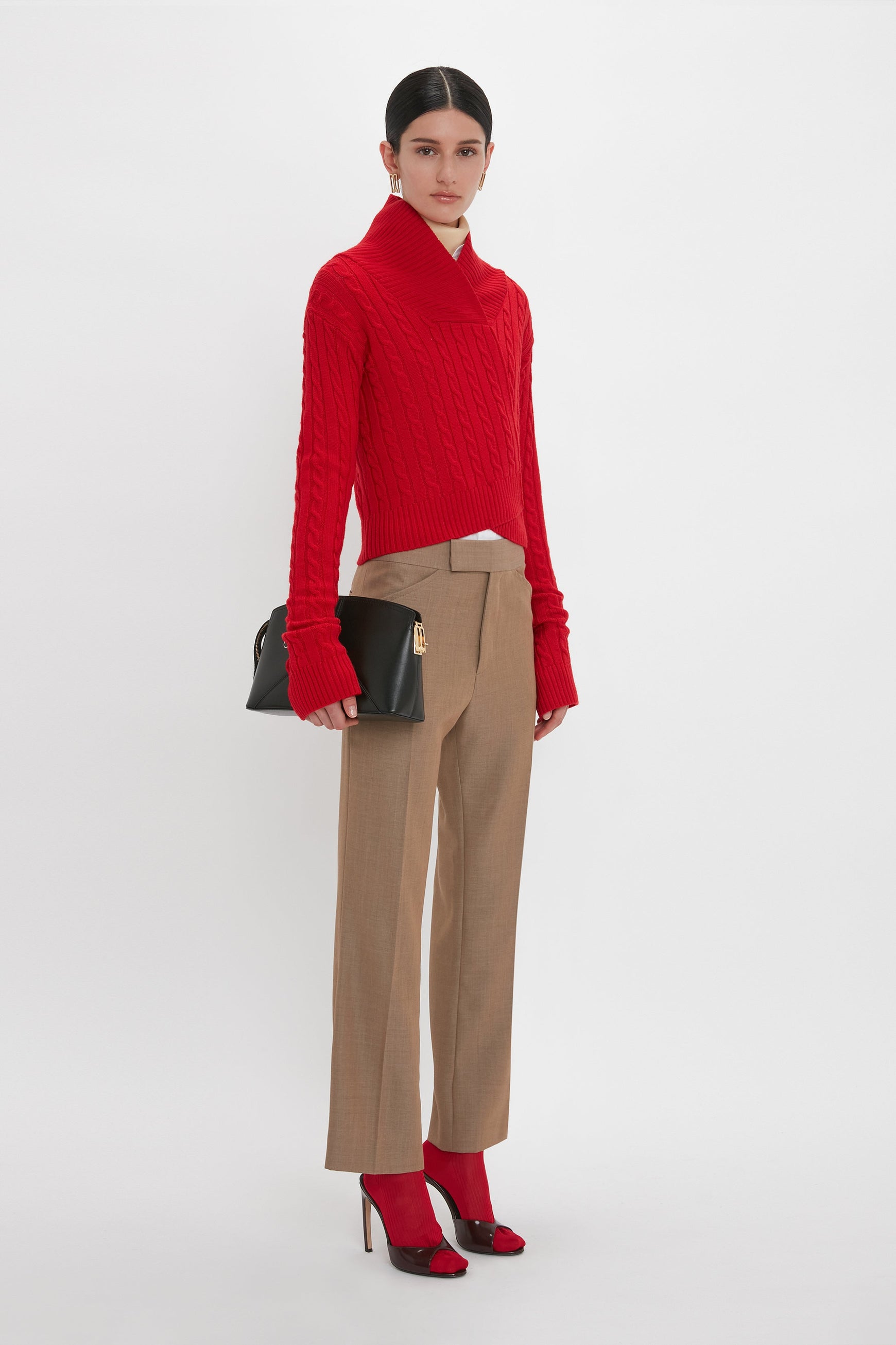 A person stands against a white background wearing a red sweater, Victoria Beckham Wide Cropped Flare Trouser In Tobacco, red shoes that reveal a flattering hint of ankle, and holding a black clutch.