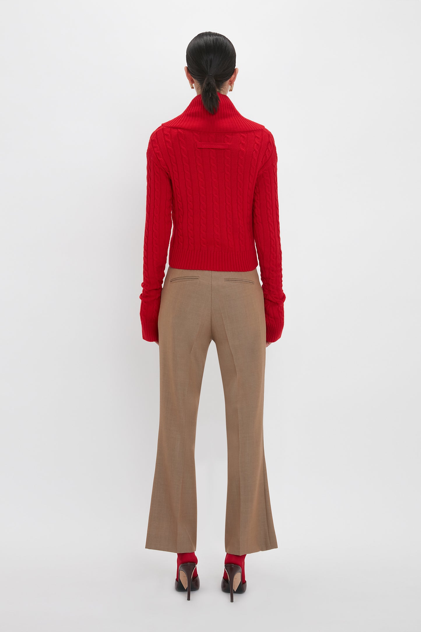 A person with dark hair in a ponytail is standing with their back to the camera, wearing a luxurious Wrap Detail Jumper In Red by Victoria Beckham, beige trousers, red socks, and black heels against a white background.