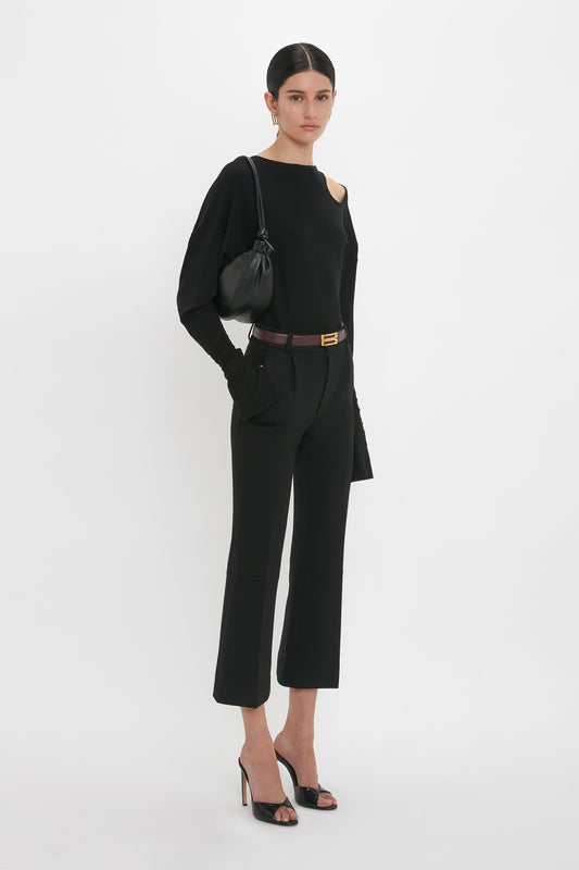 A woman modeling a black Victoria Beckham Twist Detail Jersey Top, cropped trousers, and high heels, accessorized with a belt and small bag, standing against a white background.