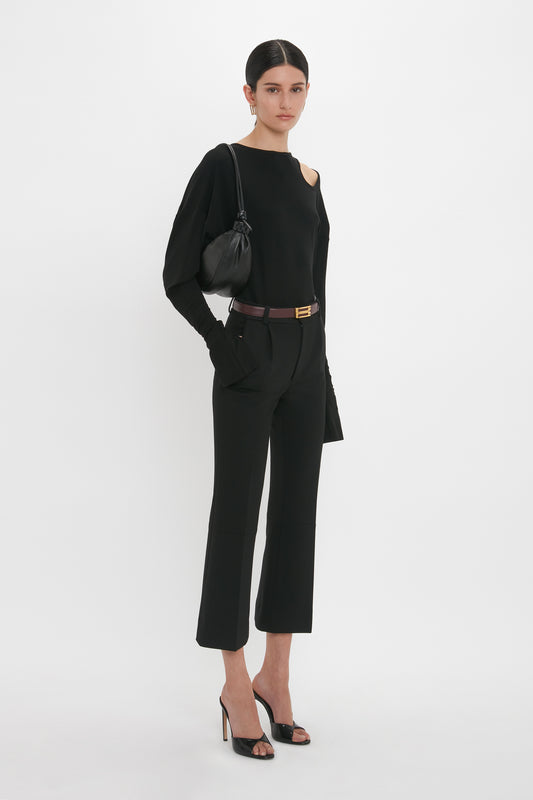 Person wearing an all-black outfit with a long-sleeve top, high-waisted pants, and the Victoria Beckham Classic Mule In Black Calf Leather featuring seductive curved heels, standing against a plain white background and holding a luxury calf leather black purse over one shoulder.