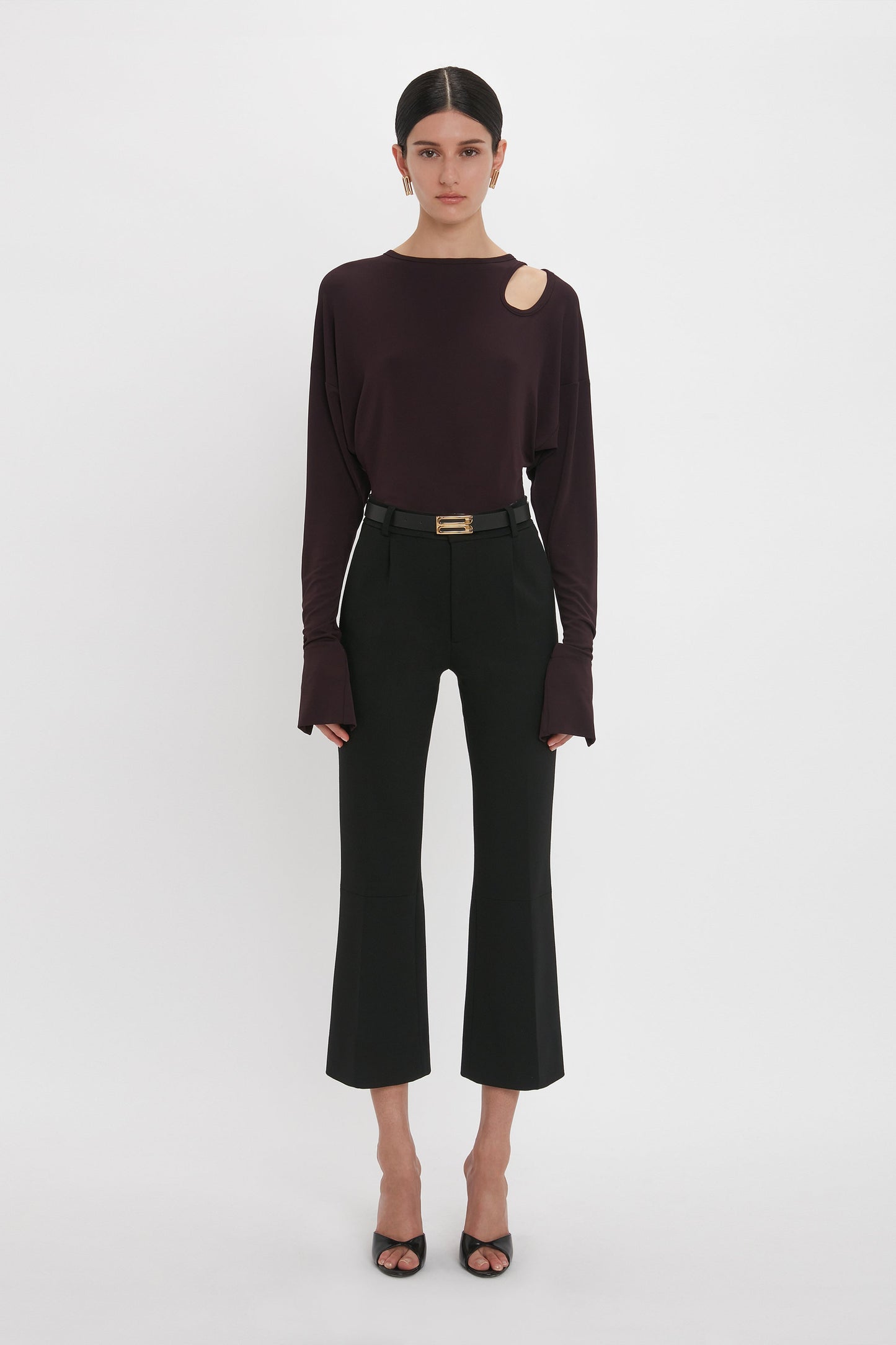 A woman stands against a white background wearing a Victoria Beckham deep mahogany twist detail jersey top with a shoulder cutout, black trousers, and black heels, accessorized with a thin belt.