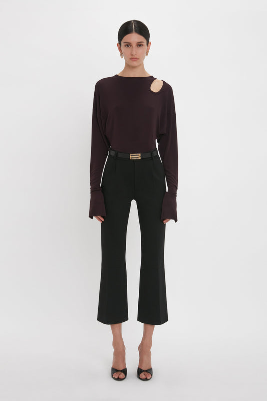 A woman stands against a white background wearing a Victoria Beckham deep mahogany twist detail jersey top with a shoulder cutout, black trousers, and black heels, accessorized with a thin belt.