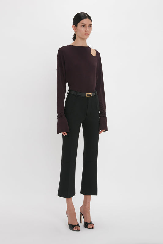 A woman in a chic burgundy Victoria Beckham Twist Detail Jersey Top in Deep Mahogany and black trousers stands against a white background, accessorized with a slim belt and black heeled sandals.