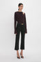 A woman in a chic burgundy Victoria Beckham Twist Detail Jersey Top in Deep Mahogany and black trousers stands against a white background, accessorized with a slim belt and black heeled sandals.