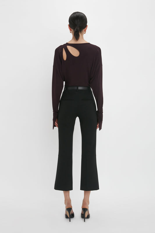 A woman stands with her back to the camera, wearing a Victoria Beckham twist detail jersey top in deep mahogany and black flared trousers, against a white background.