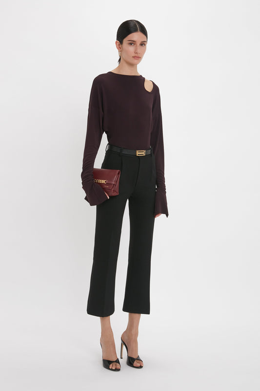 Woman in a stylish outfit, featuring a Victoria Beckham Twist Detail Jersey Top in Deep Mahogany, black trousers, and a small clutch, posing against a plain white background.