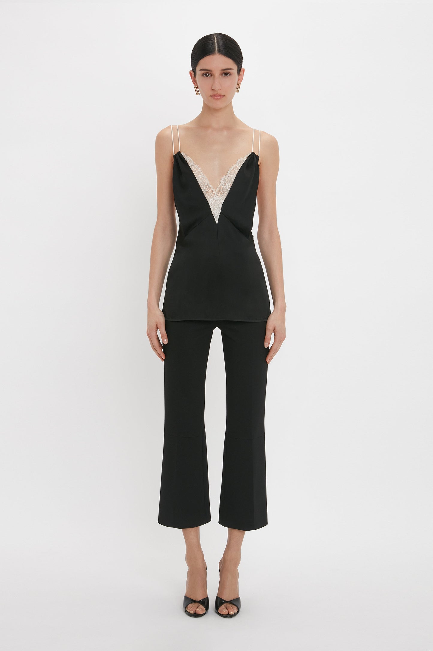 A person stands against a plain white background wearing a black Lace Detail Cami Top In Black by Victoria Beckham with a deep-V neckline and black flare pants, paired with black open-toe shoes.