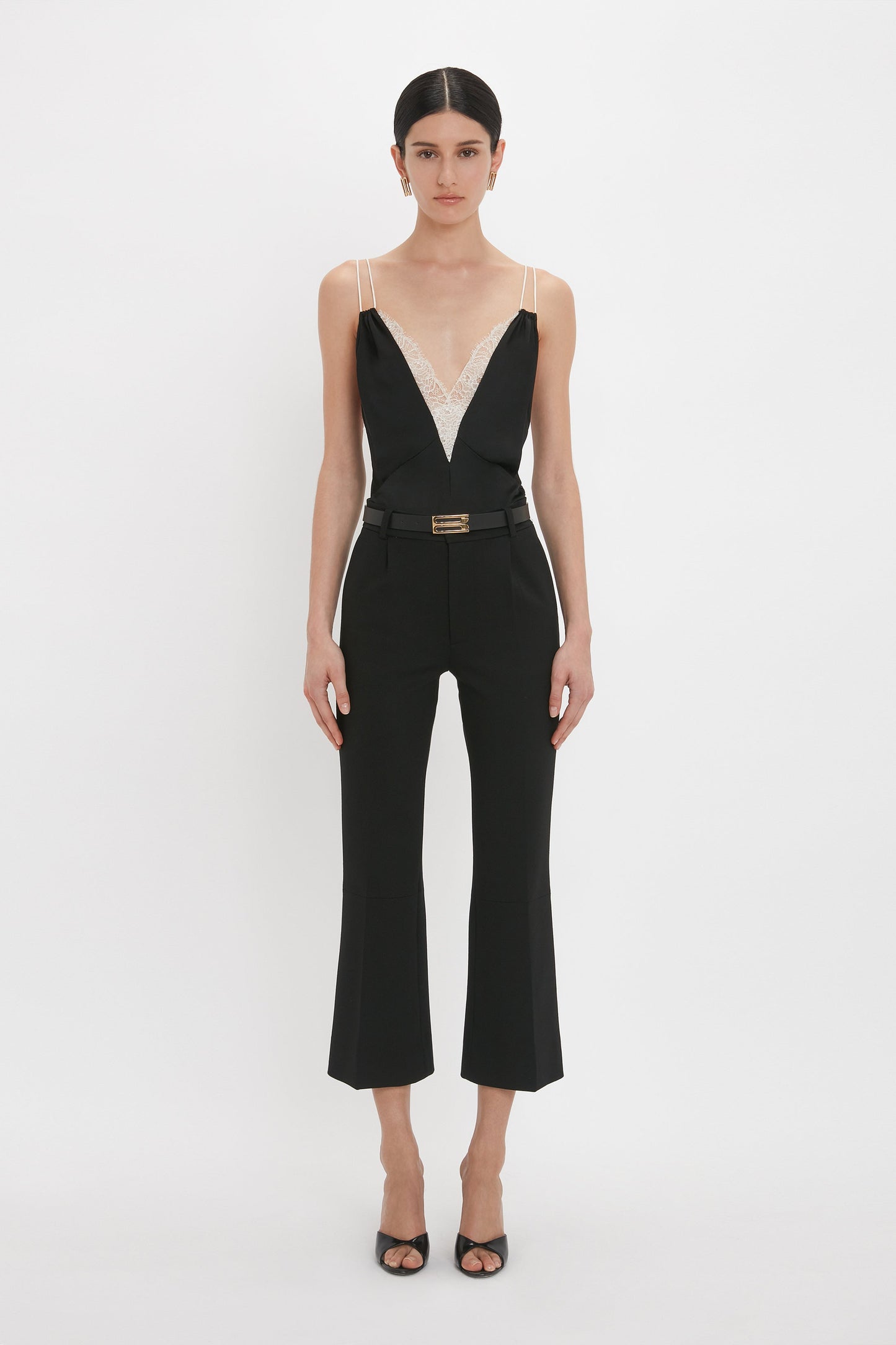 A person stands against a plain white background, wearing a black Victoria Beckham Lace Detail Cami Top In Black with a plunging neckline and lace trim, cropped pants, and black heeled sandals.