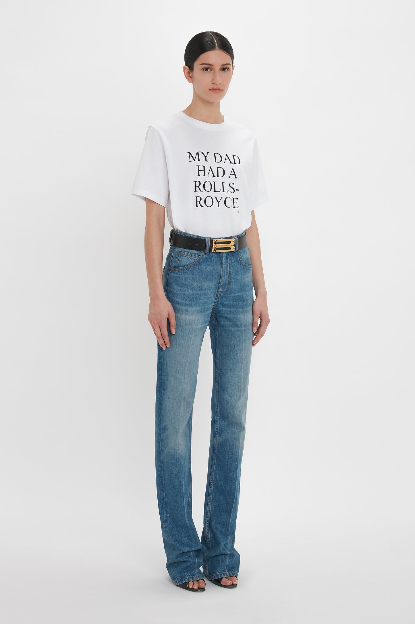 A woman stands against a white background wearing a white T-shirt that reads "MY DAD HAD A ROLLS-ROYCE," Julia Jean In Broken Vintage Wash by Victoria Beckham, and black open-toe shoes. She has dark hair pulled back, a black belt with a gold buckle, and her high waistline jeans are made from 100% cotton denim.