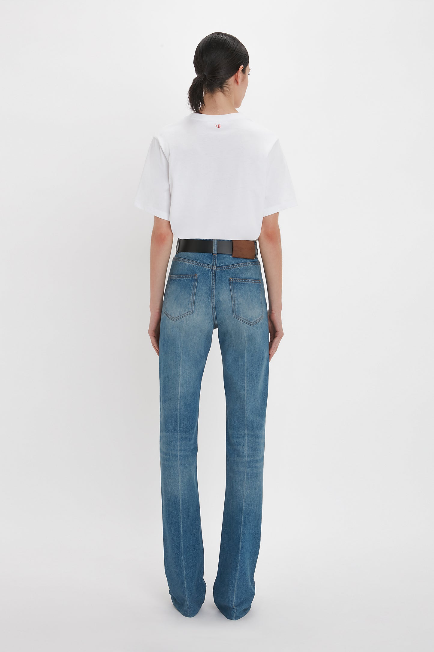 A person with dark hair is seen from the back, wearing a white short-sleeve shirt and blue distressed Julia Jean In Broken Vintage Wash by Victoria Beckham made from 100% cotton denim. They are standing against a plain white background.