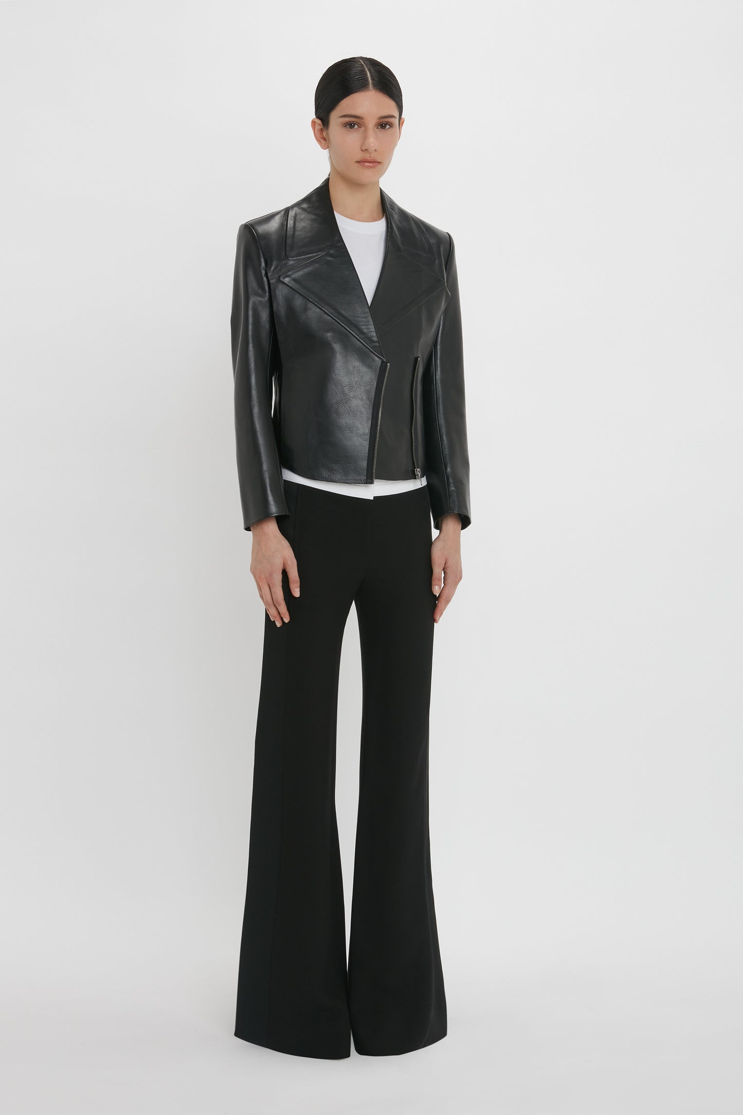 A person stands against a plain white background, dressed in a Victoria Beckham Tailored Leather Biker Jacket In Black, white shirt, and black flared pants.