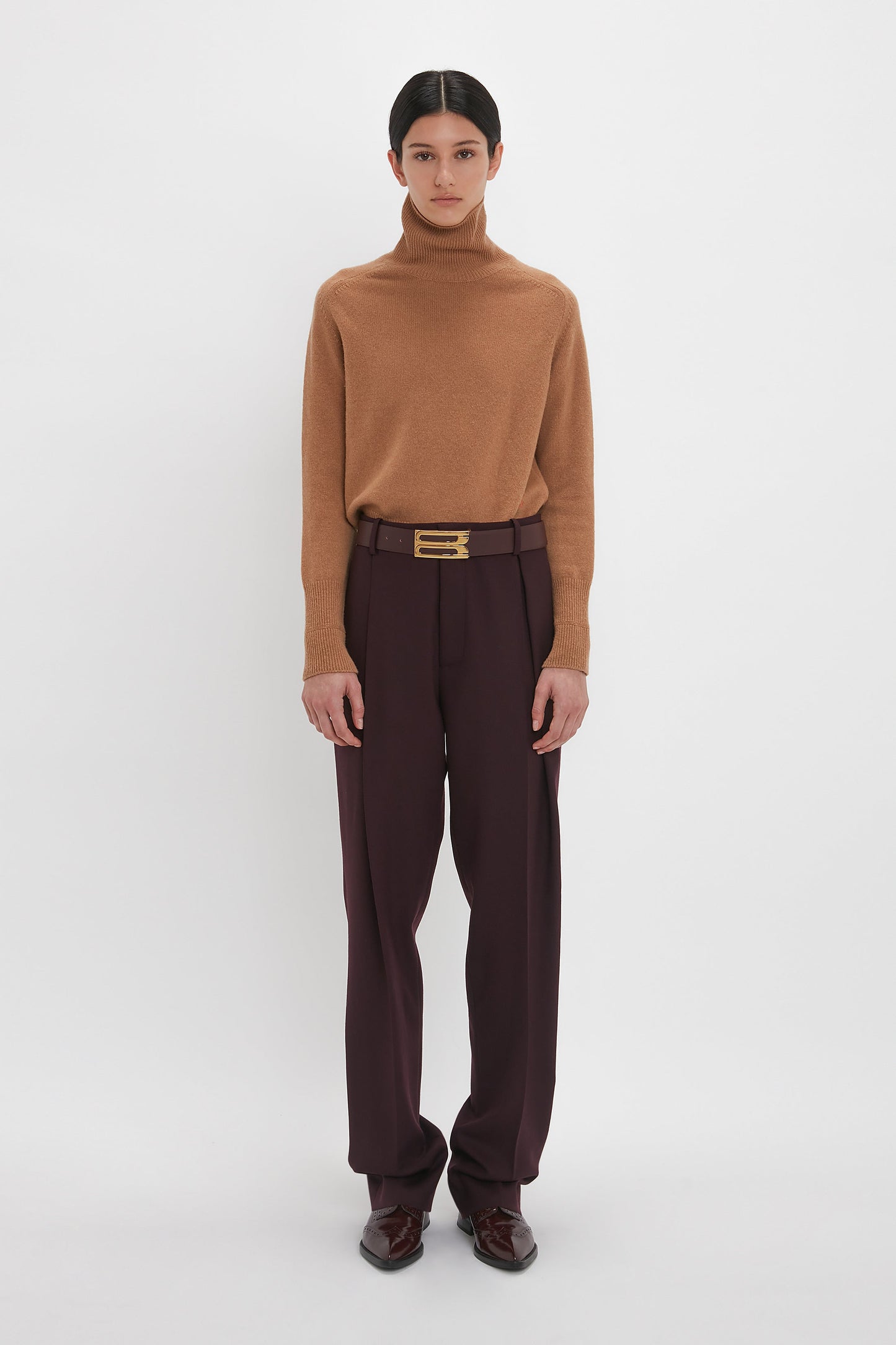 Person standing against a white background, wearing a brown turtleneck sweater, Victoria Beckham Asymmetric Chino Trouser In Deep Mahogany with a narrow leg silhouette and a gold buckle belt, and dark shoes.