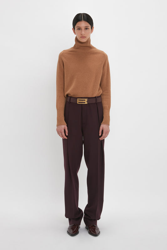 A person wearing a Victoria Beckham lambswool polo neck jumper in tobacco and dark trousers with a black belt, standing in front of a plain white background.