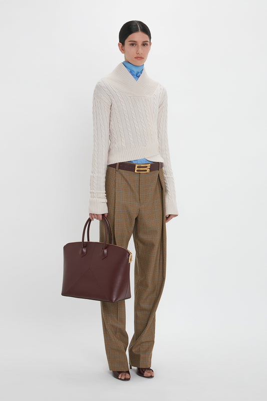 Person standing against a plain background wearing a cream sweater, blue shirt, brown plaid pants, and holding a Victoria Bag In Burgundy Leather by Victoria Beckham with an adjustable strap.