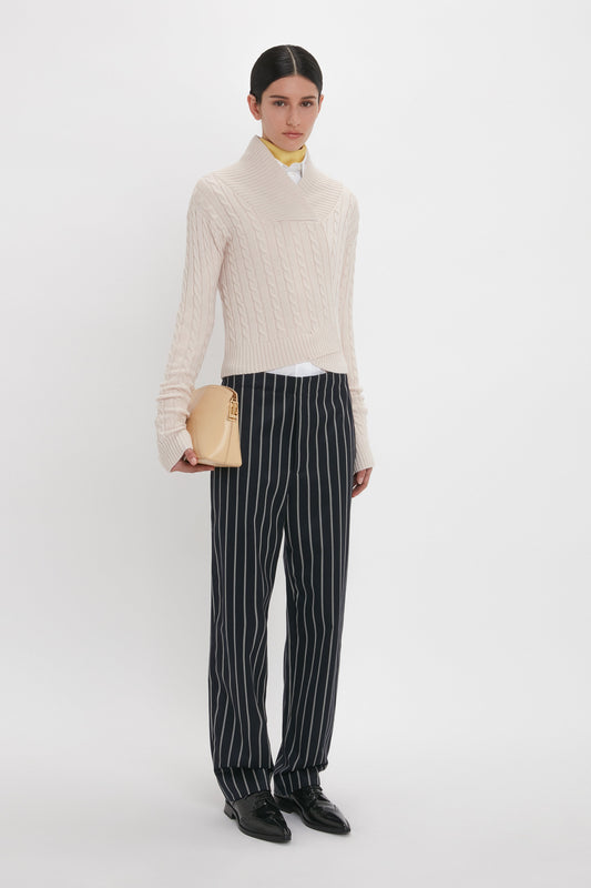 A person stands against a plain background, dressed in a white cable-knit sweater, Tapered Leg Trouser In Midnight-White by Victoria Beckham, black shoes, and holding a beige clutch bag.