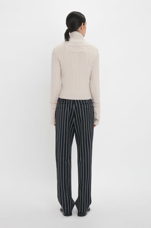 A person wearing a beige cable-knit turtleneck sweater and Victoria Beckham Tapered Leg Trouser In Midnight-White stands facing away from the camera against a white background.