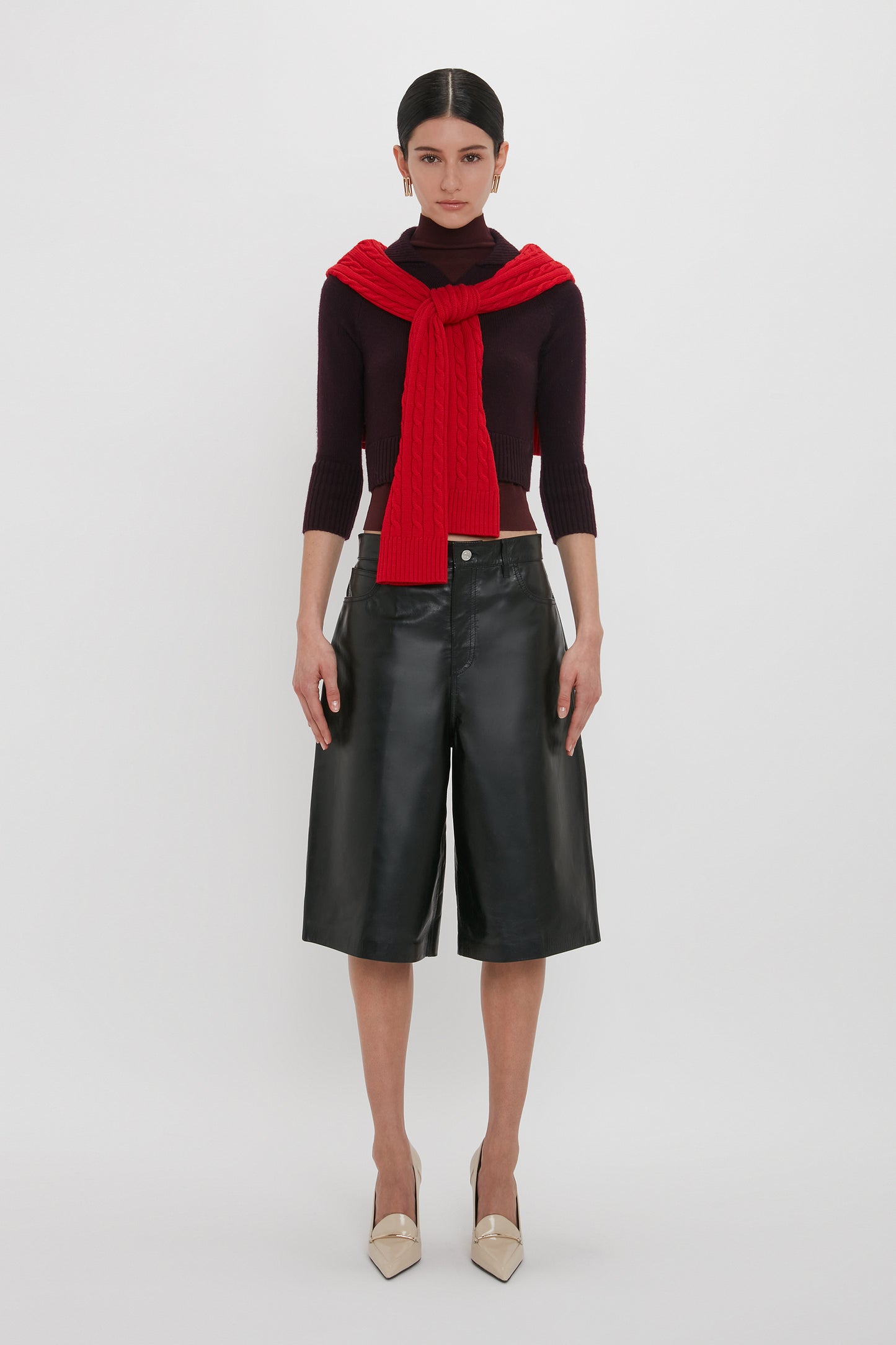 A person stands against a white background, wearing a dark turtleneck with a red sweater draped over the shoulders, **Victoria Beckham Leather Bermuda Short In Black** featuring five-pocket detailing, and beige shoes.