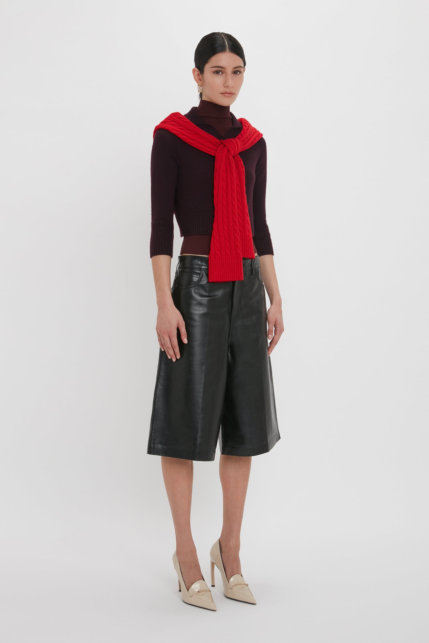 A woman stands against a plain background wearing a cropped black sweater, a red sweater draped over her shoulders, Victoria Beckham Leather Bermuda Short In Black with five-pocket detailing, and beige pointed high heels.