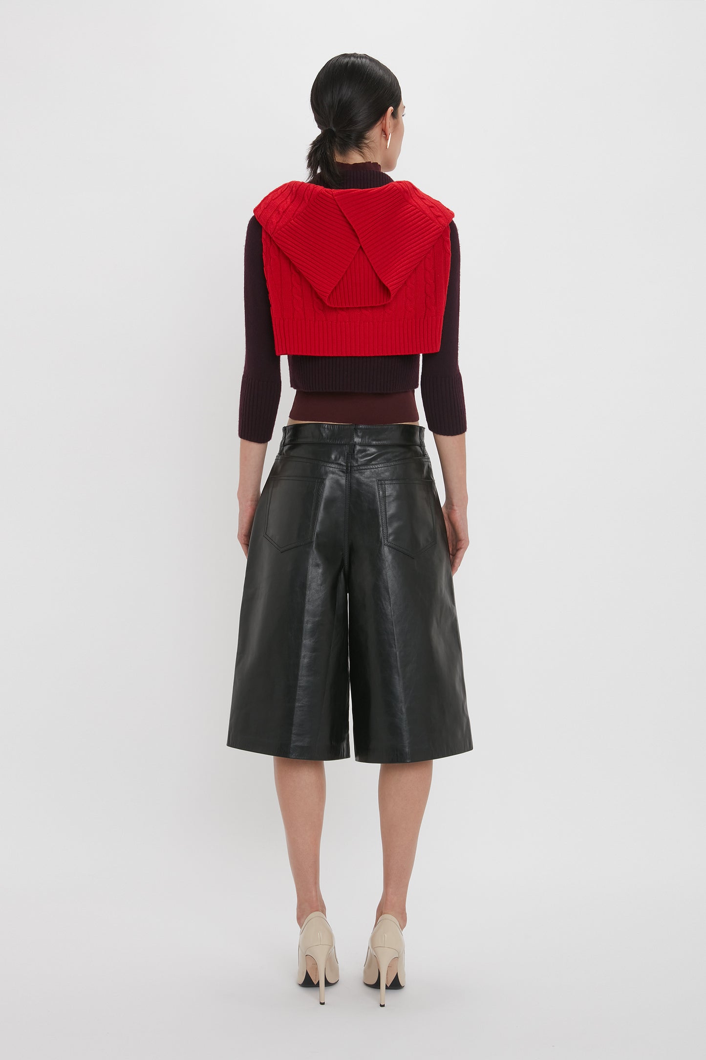 A person is standing with their back to the camera, wearing a red sweater draped over their shoulders, a dark long-sleeve top, Victoria Beckham Leather Bermuda Short In Black with five-pocket detailing, and beige high heels.