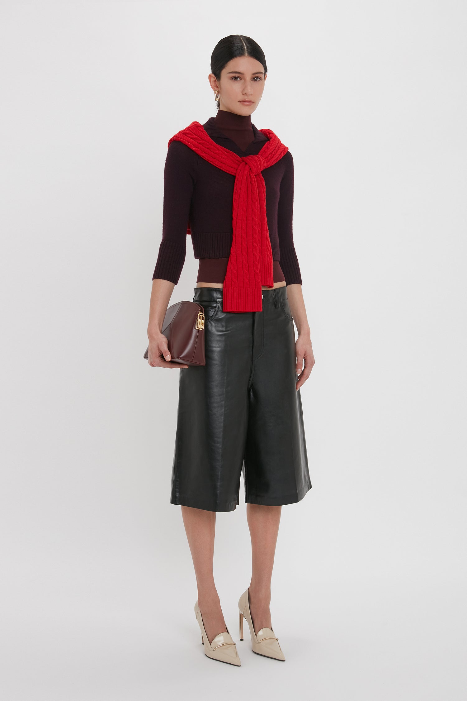 Woman wearing a black top with a red sweater draped over her shoulders, Leather Bermuda Short In Black by Victoria Beckham featuring five-pocket detailing, beige heels, and holding a maroon clutch against a plain white background.