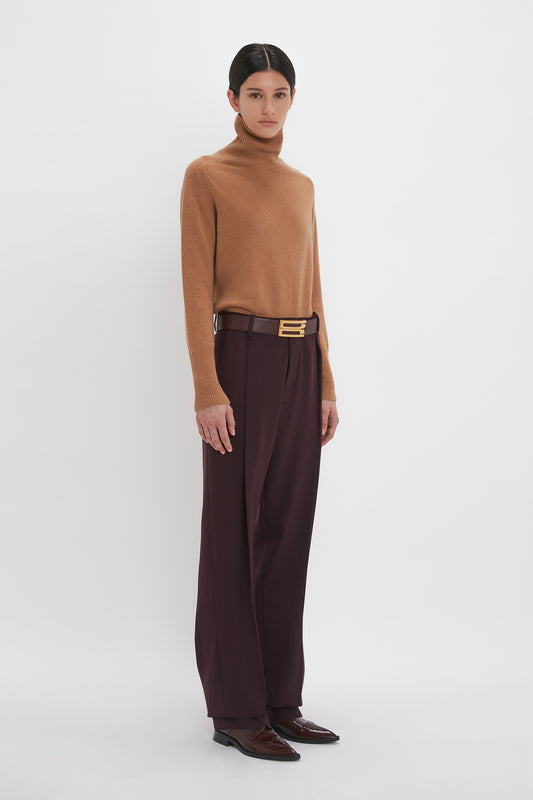 A woman stands in a neutral pose wearing a Victoria Beckham lambswool polo neck jumper in tobacco, dark wide-leg trousers, and brown loafers against a plain white background.