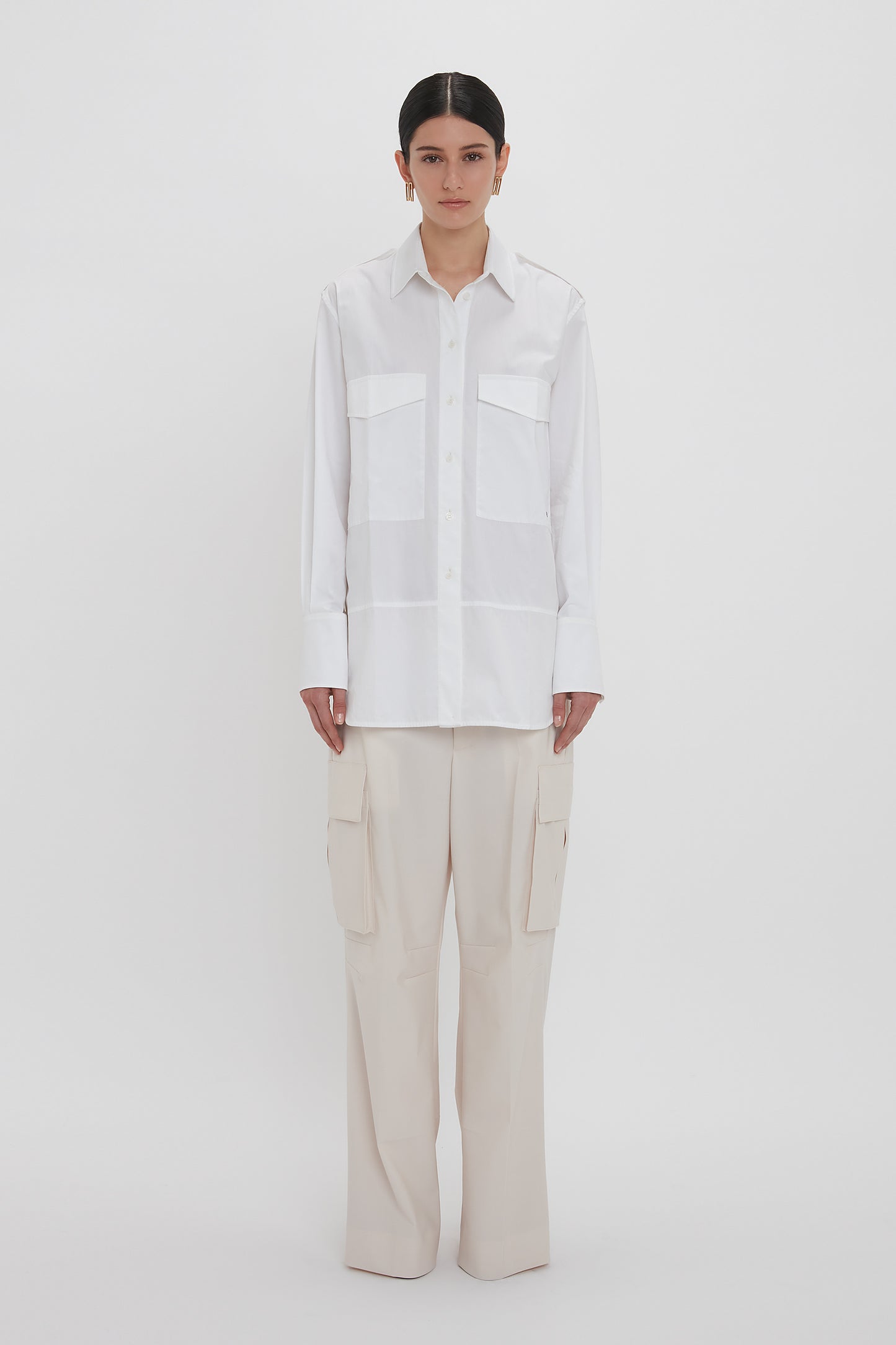 A person stands against a white background wearing an Oversized Pocket Shirt In White by Victoria Beckham made of organic cotton and light beige Relaxed Cargo Trouser.