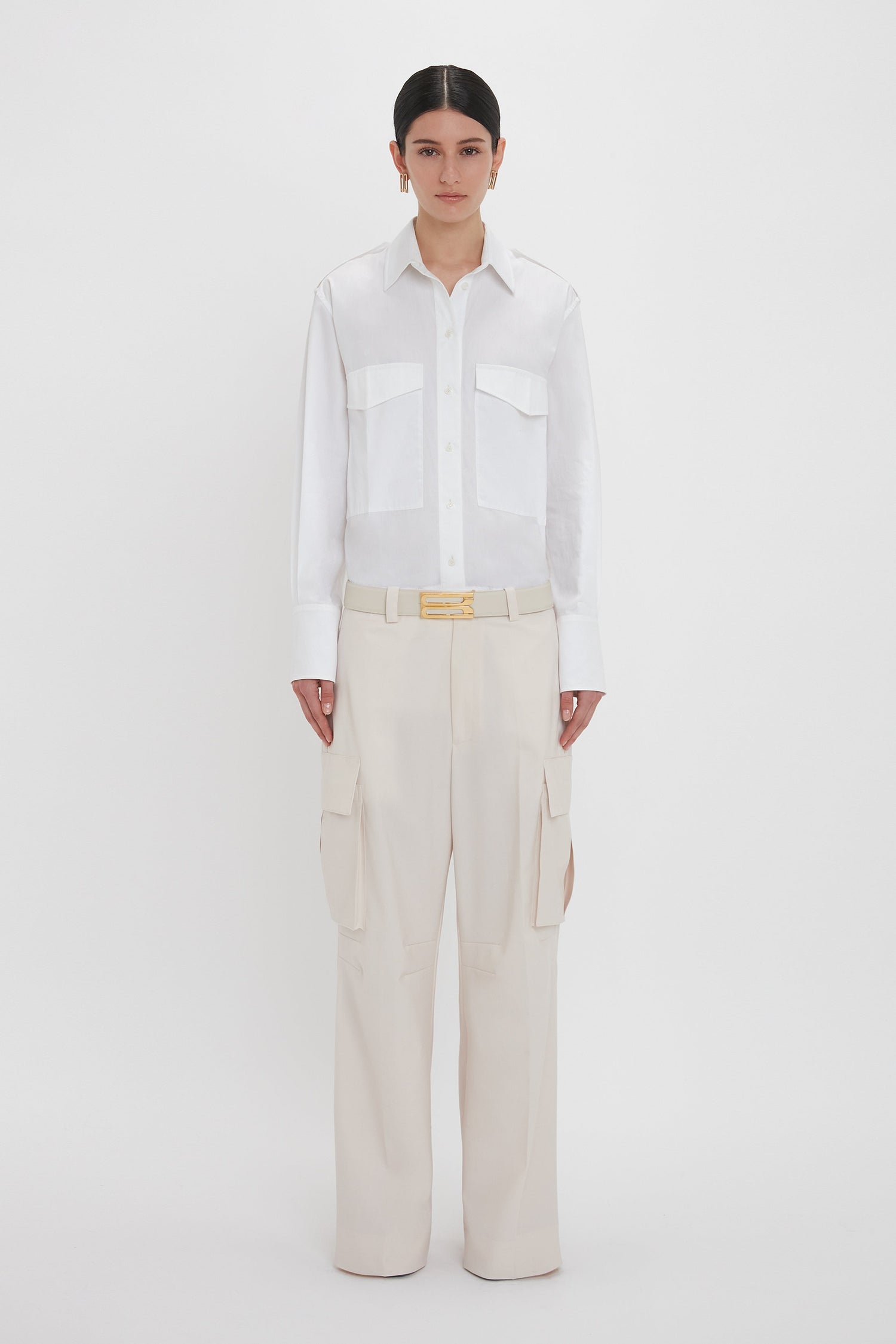 A person stands against a plain white background wearing an Oversized Pocket Shirt In White by Victoria Beckham made from organic cotton and cream-colored Relaxed Cargo Trousers featuring a gold belt buckle.