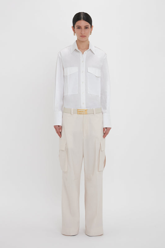 A person stands against a plain white background, wearing a white button-up shirt and light beige, military-inspired Relaxed Cargo Trouser In Bone from Victoria Beckham, accented with a gold belt buckle.