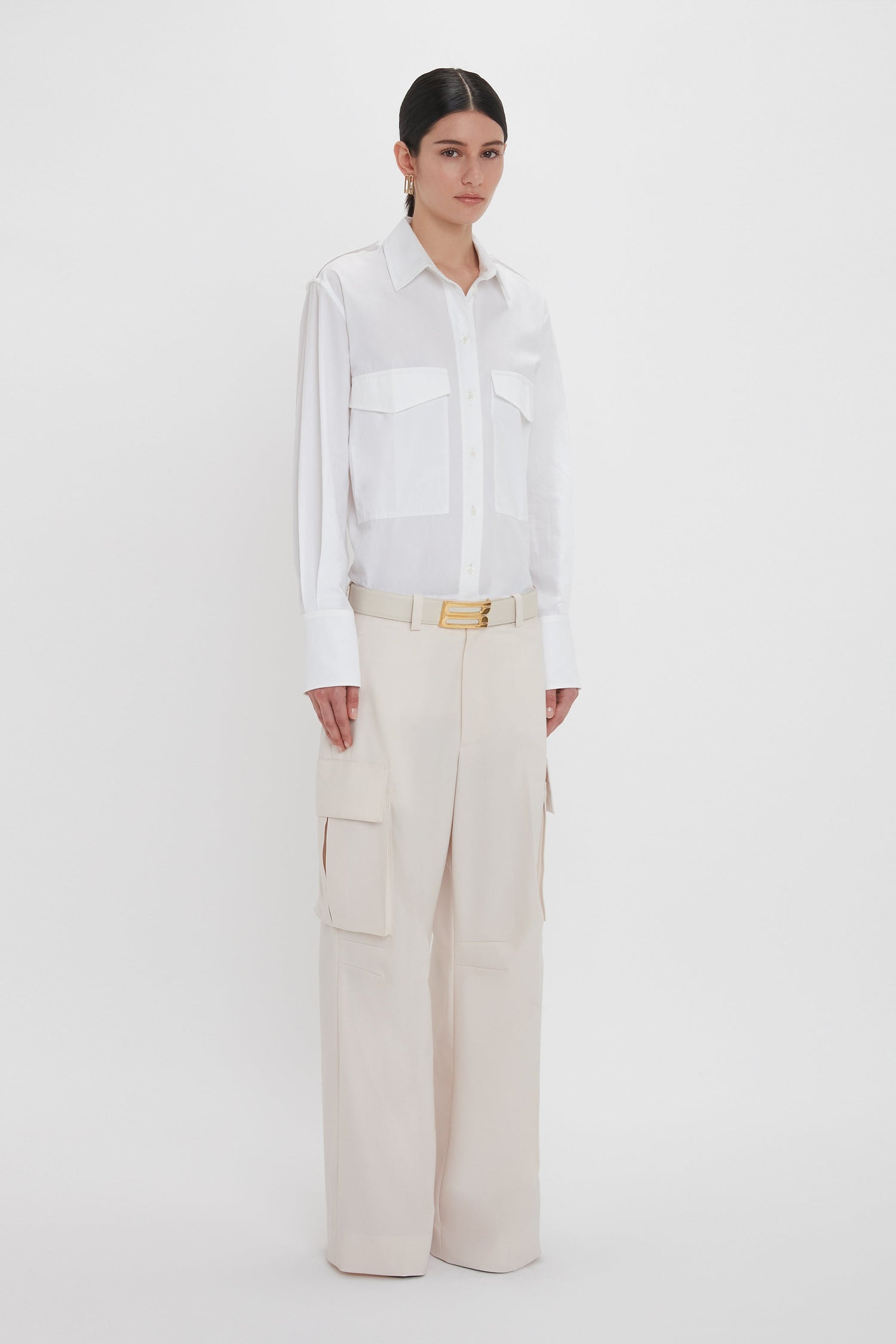 A person stands against a plain white background, wearing a Victoria Beckham Oversized Pocket Shirt In White and beige relaxed cargo trousers with a belt. They have dark hair pulled back and are looking slightly off-camera.