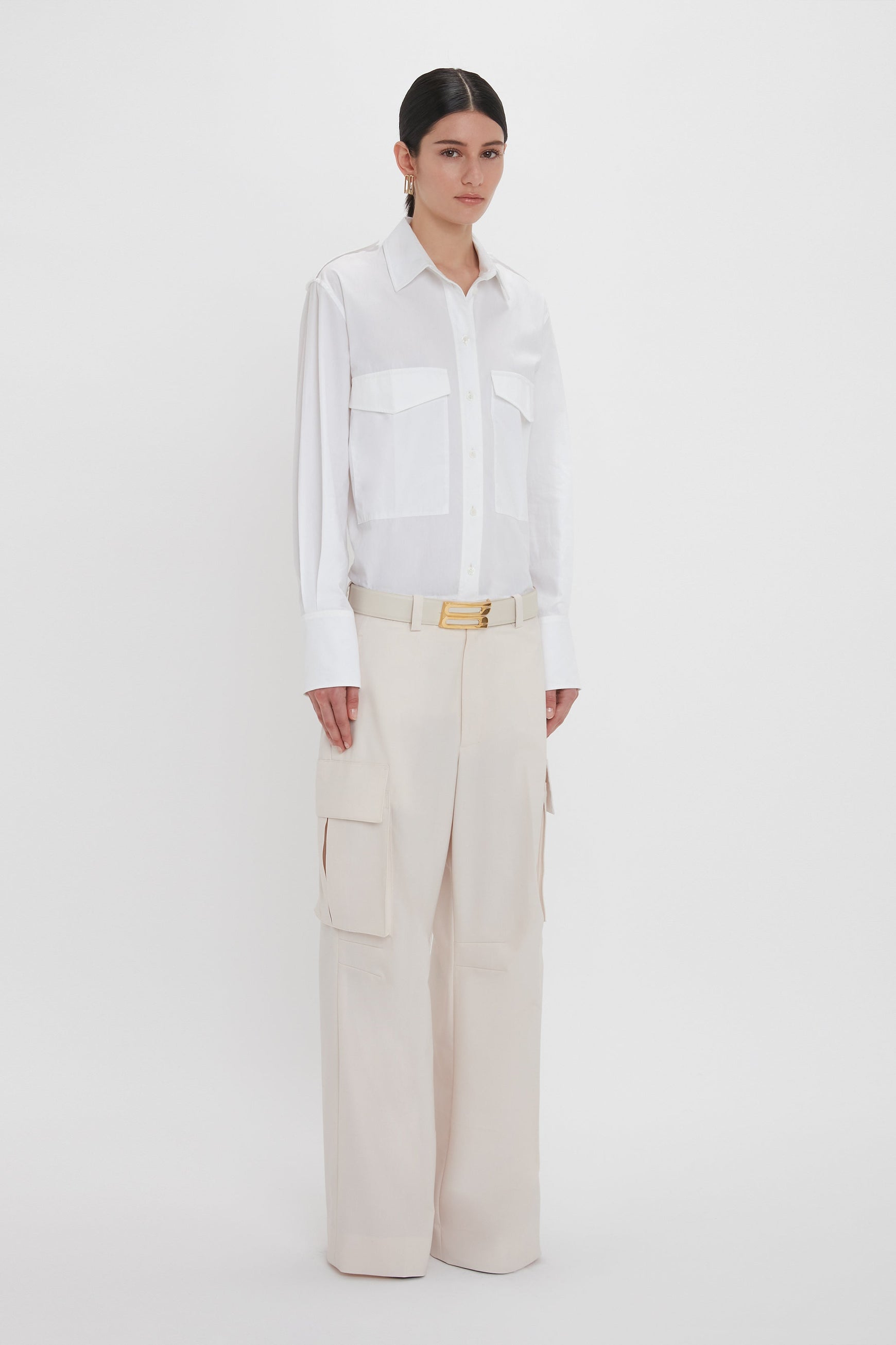 A person stands against a white background wearing a white button-up shirt and Victoria Beckham's Relaxed Cargo Trouser In Bone. Their hands rest by their sides.