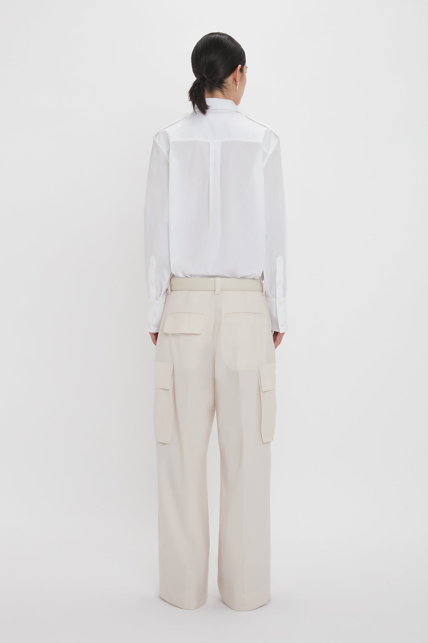 A person with dark hair wearing a white Victoria Beckham Oversized Pocket Shirt In White and cream-colored Relaxed Cargo Trouser stands facing away in a minimalistic setting.