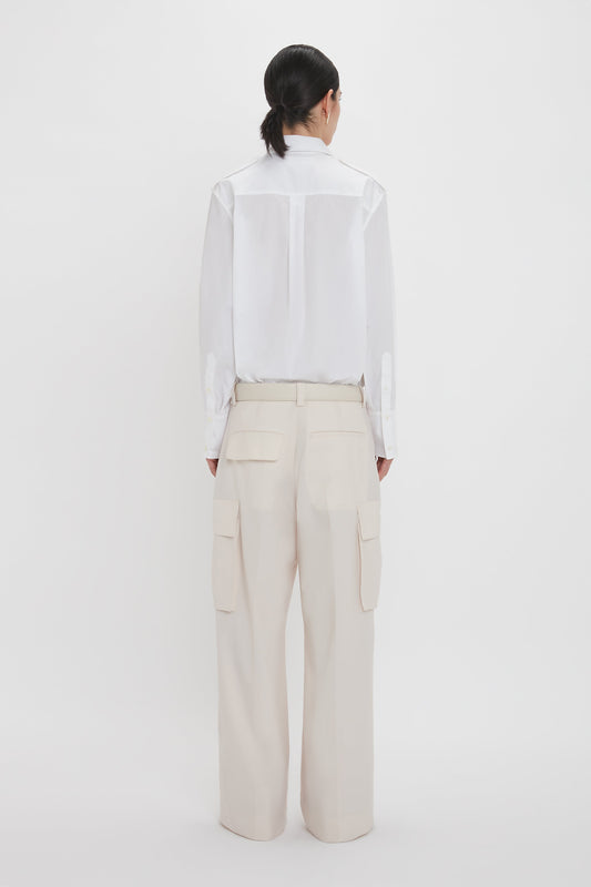 Person standing against a white background, wearing a white shirt and the Relaxed Cargo Trouser In Bone by Victoria Beckham, seen from behind. The 100% cotton material and military-inspired design add a rugged touch to the outfit.