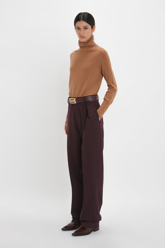 A woman stands against a white background, wearing a Victoria Beckham camel lambswool polo neck jumper in tobacco, brown belted trousers, and dark loafers.