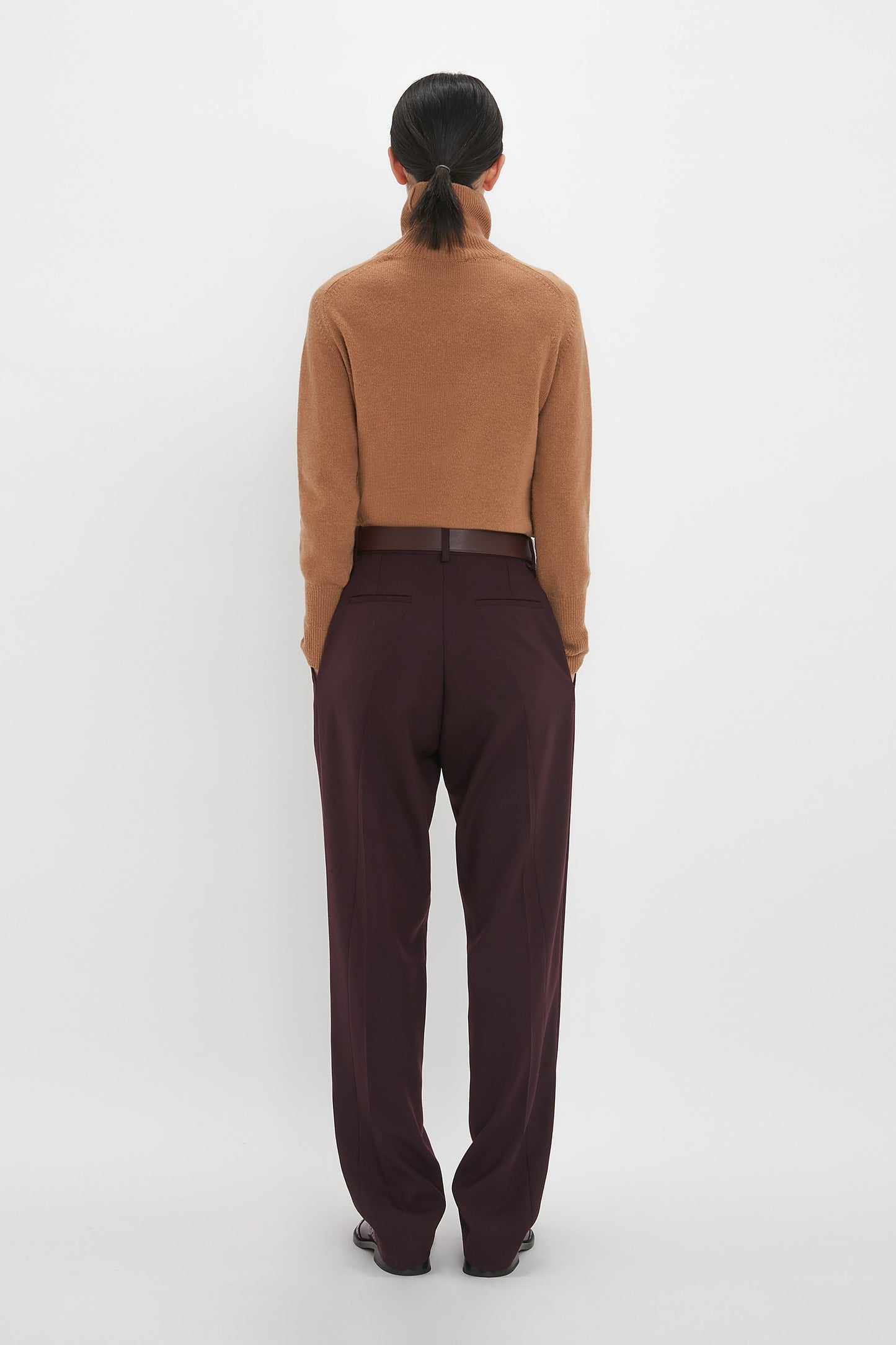 A person stands with their back to the camera, wearing a brown long-sleeve sweater and high-waisted, dark brown Asymmetric Chino Trouser In Deep Mahogany by Victoria Beckham. Their hair is tied back in a low ponytail, highlighting the narrow leg silhouette of their recycled wool pants against the plain white background.