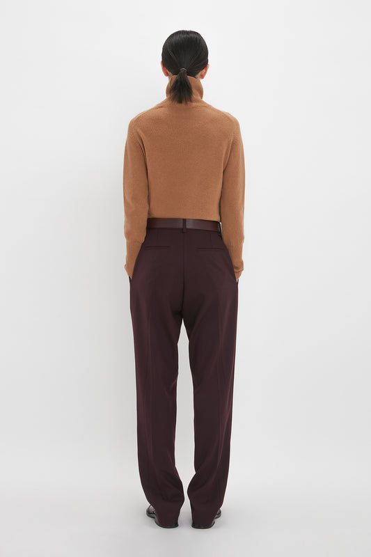 Woman viewed from behind, wearing a Victoria Beckham lambswool polo neck sweater in tobacco and dark maroon trousers, standing against a white background.