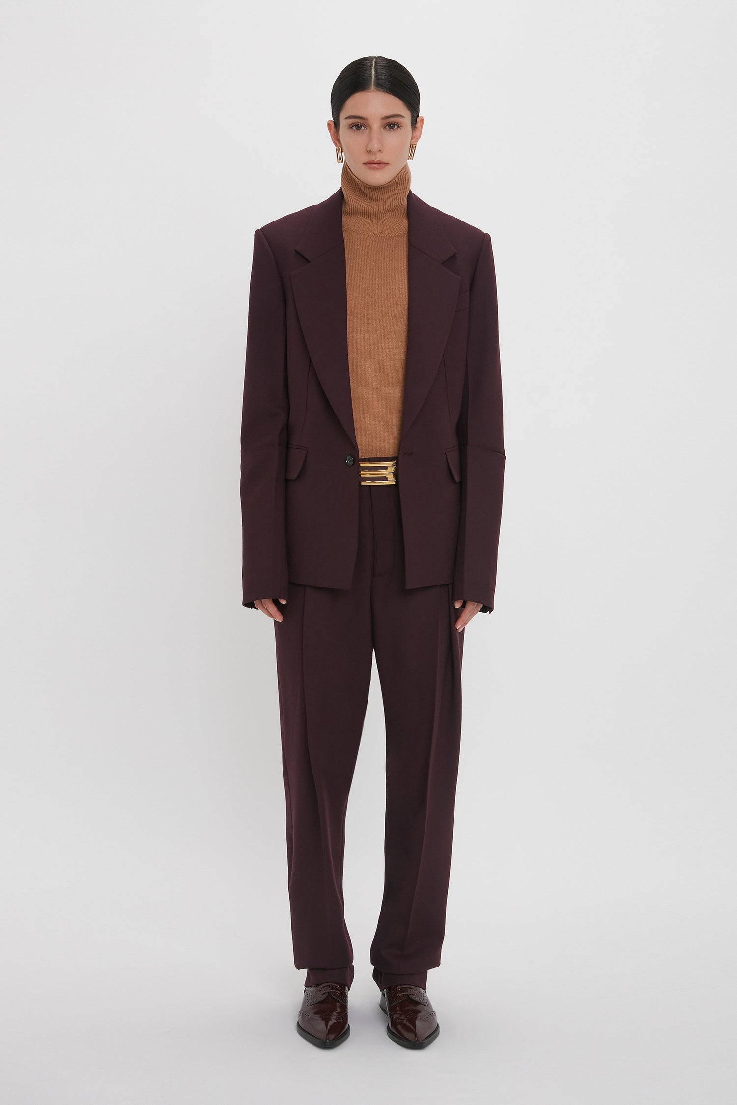 Person standing against a white background, dressed in a Victoria Beckham Sleeve Detail Patch Pocket Jacket In Deep Mahogany and pants with a brown turtleneck underneath, wearing dark shoes and gold earrings.