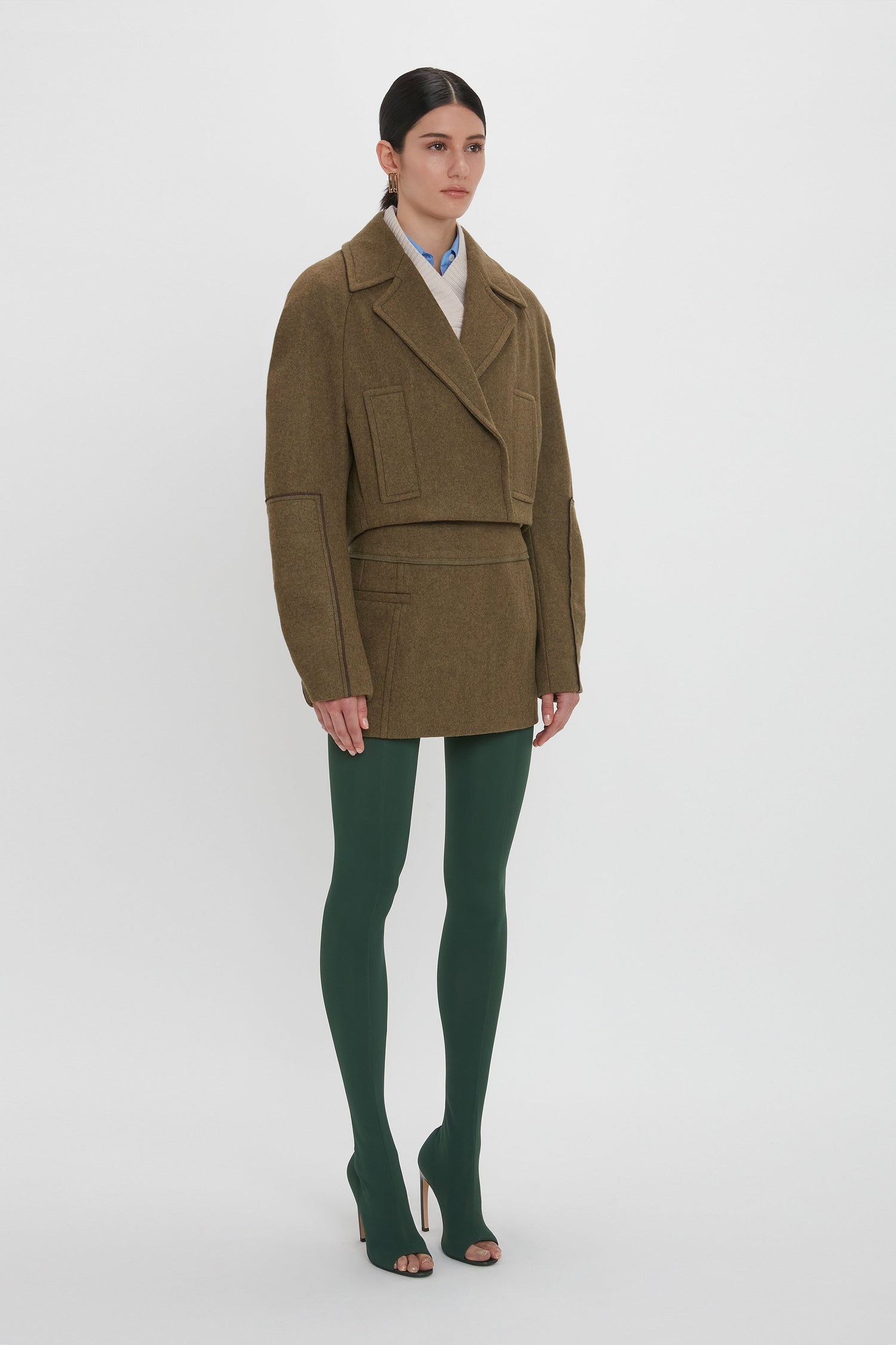 A person stands facing slightly to the right, wearing a waist-defining Cropped Pea Coat In Khaki by Victoria Beckham, matching skirt, green tights, and beige high heels against a plain white background.