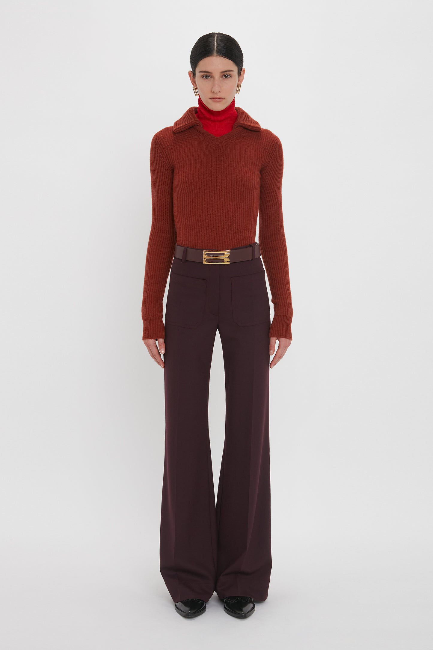 A person stands against a white background wearing a Double Collared Jumper In Russet by Victoria Beckham, dark wide-leg pants, a red turtleneck underneath, and black shoes.